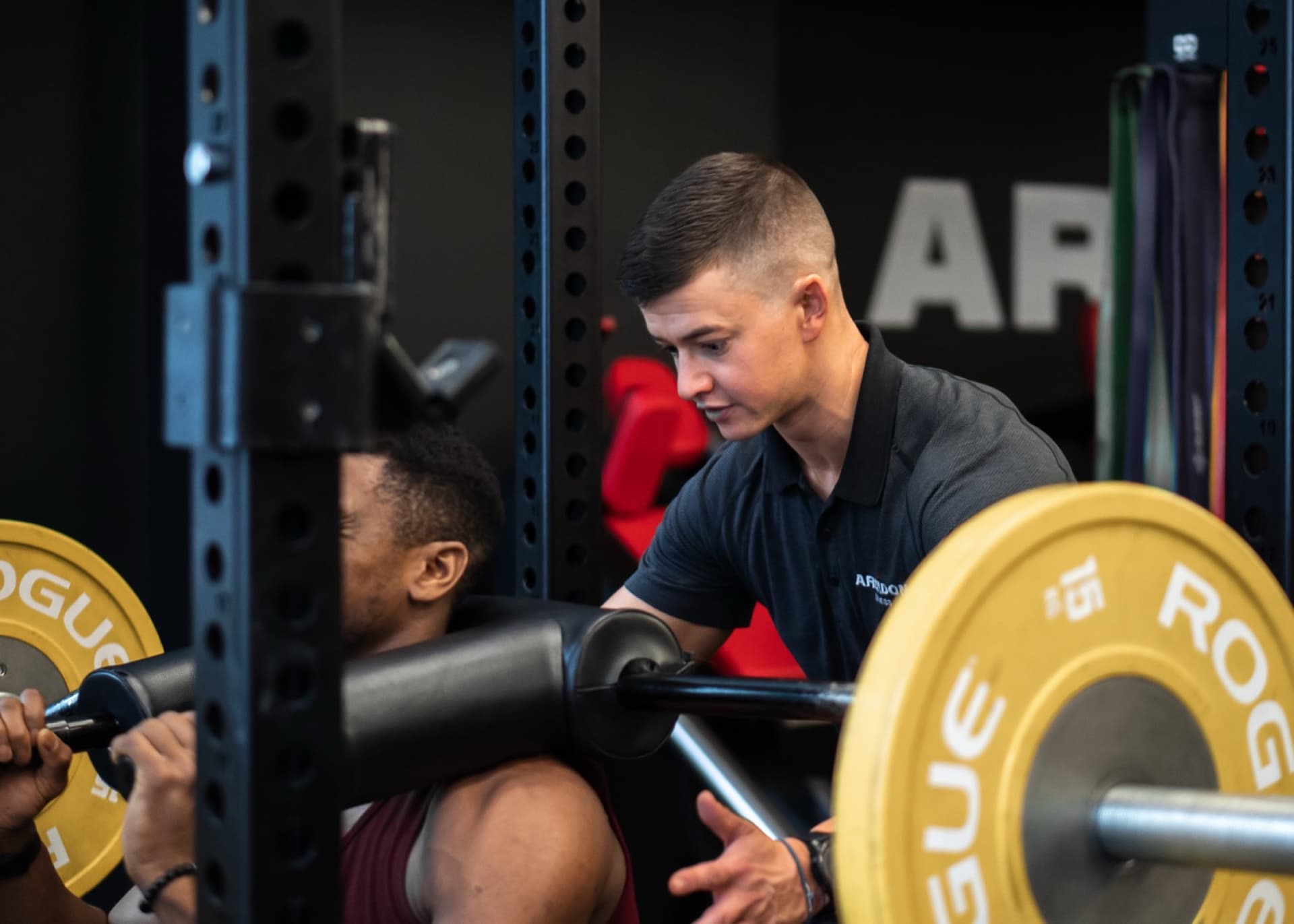 Arzadon trainer in branded polo spotting male client squatting under Rogue rack at Arzadon Fitness Toronto